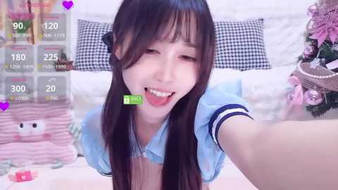 A video of an Asian woman with long dark hair, wearing a light blue top, playfully sticking out her tongue, surrounded by virtual camera features, in a cozy bedroom.