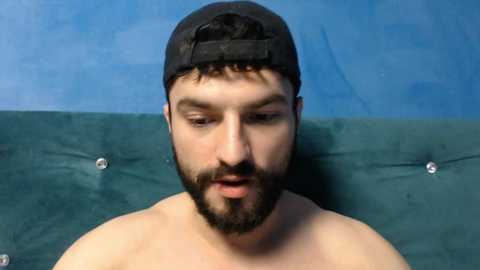 Video of a bearded man with short dark hair wearing a black baseball cap, sitting shirtless against a blue wall with metal studs.