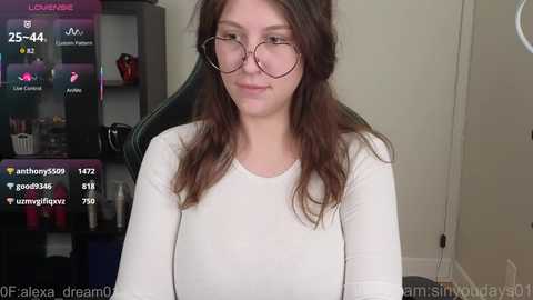 Video of a young woman with long brown hair, wearing glasses, a white top, and sitting on a black gaming chair. The background includes a shelf with various items and a screen displaying live stream details.