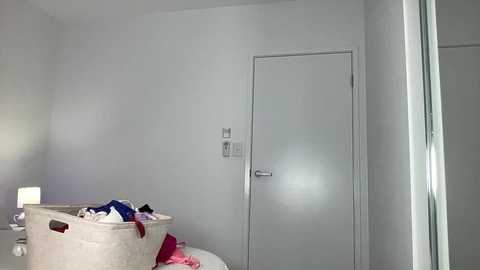 A video of a minimalist, white-walled bedroom with a beige laundry basket overflowing with clothes, a door with a silver handle, and a small bedside table with a lamp.