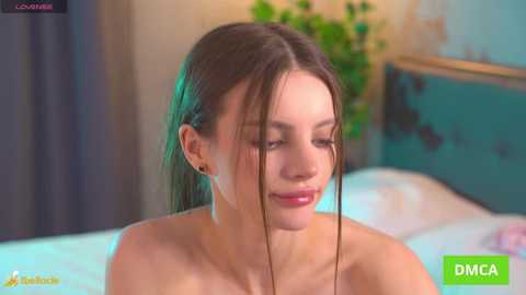 A video of a young, topless woman with wet hair, wearing a pink necklace, smiling softly, in a dimly-lit bedroom with a green wall and bed.