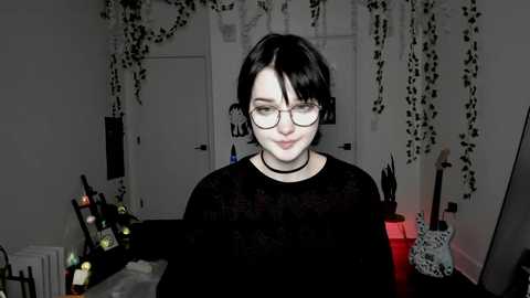 A video of a pale-skinned woman with short black hair, glasses, and a black choker, standing in a dimly lit, cluttered room with wine bottles, a guitar, and hanging plants.