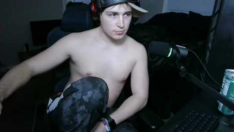 Media: A young, shirtless, fair-skinned man with a small build, wearing headphones, sits in a dark room with a computer setup. He has a contemplative expression, and the room is dimly lit with a cluttered background.