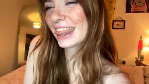 Video of a smiling young woman with fair skin, long brown hair, and freckles, wearing a nose ring, in a cozy bedroom with warm lighting, framed art, and a bedside lamp.