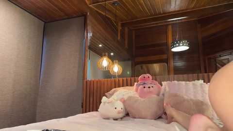 A video of a cozy, wooden-paneled room with a plush, pink pig plush toy on a bed, surrounded by pastel-colored pillows and a white duvet.