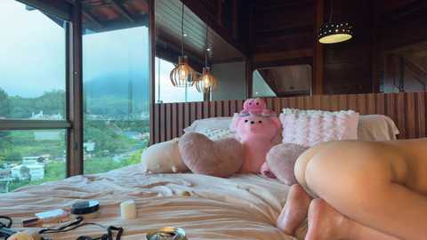 Video of a cozy, rustic bedroom with wooden walls and ceiling, featuring a plush pink piggy bank, fluffy pillows, and a view of a lush green landscape through large windows.