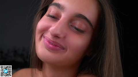 Media: Video of a young woman with light brown skin and long dark hair, smiling gently, wearing minimal makeup. The background is dark, emphasizing her face.