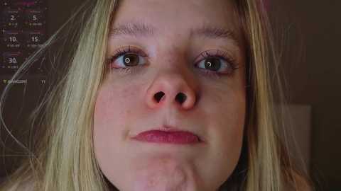 Media: A close-up video of a blonde woman with fair skin, green eyes, and a pink nose. She has a neutral expression, looking directly at the camera. The background is blurry, showing a dark, textured surface.