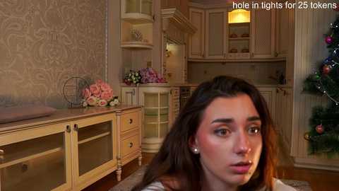 Media: Video of a young woman with long brown hair, wearing makeup, sitting in a cozy, vintage kitchen with cream-colored cabinets, a Christmas tree, and a hole-in-light for 25tokyokern.