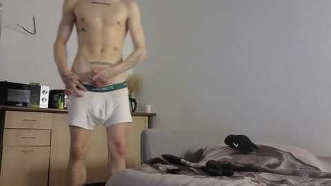 Video of a shirtless, slender, light-skinned man in white boxer briefs, standing in a dimly lit bedroom with a bed, dresser, and microwave.