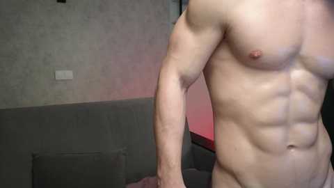 Video of a muscular, light-skinned man from the waist up, posing naked in a dimly lit room with gray walls and a red light. His abs and chest are prominently displayed, highlighting his physique.