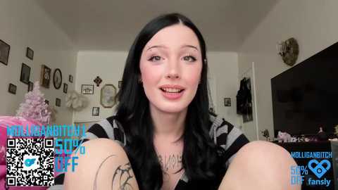 A video of a smiling, pale-skinned woman with long black hair, wearing a black and white striped top, in a cluttered, white-walled room. Text overlays include \"MILFANUTTY\" and QR codes.