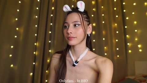 Video of a young, slender, topless Asian woman with long brown hair, wearing bunny ears, a white choker, and a pendant necklace. She stands against a backdrop of golden fairy lights.