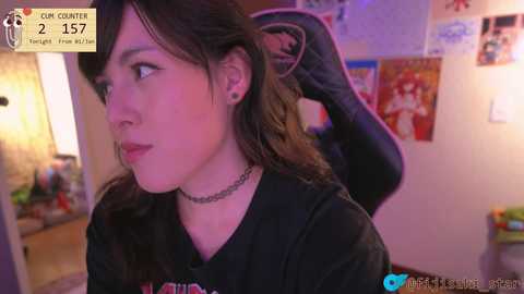 A video of a young woman with pale skin, dark hair, and a black choker, wearing a black shirt, standing in a bedroom with posters on the wall.