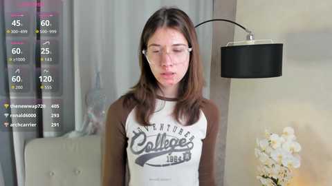 Media: Video of a young woman with straight brown hair, wearing white earbuds and a \"Colleges of the World\" shirt, standing in a modern, softly lit room with a black lampshade.