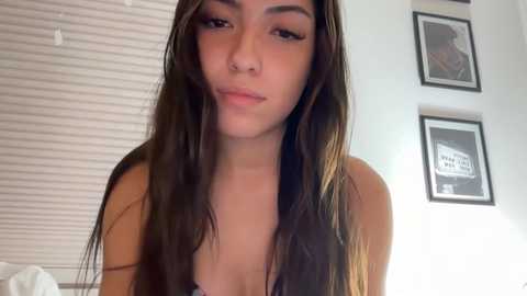 Video of a young woman with long brown hair and light skin, wearing a black top, standing indoors near white blinds and framed art on a white wall.