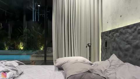 Video of a modern bedroom with a sleek, gray upholstered headboard, white sheets, and a tall, sheer white curtain. The background shows a large glass window with a pool visible outside. The room has a minimalist, contemporary style.