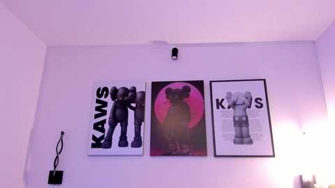 A video of a minimalist room with white walls, featuring three framed artworks: two black-and-white prints of Kanye West and one of the iconic KAWS character, all illuminated by a single hanging lamp.