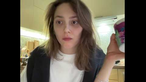 Video of a young woman with shoulder-length brown hair, fair skin, and a white T-shirt, holding a colorful ceramic mug, standing in a modern kitchen with beige cabinets and a tiled backsplash.