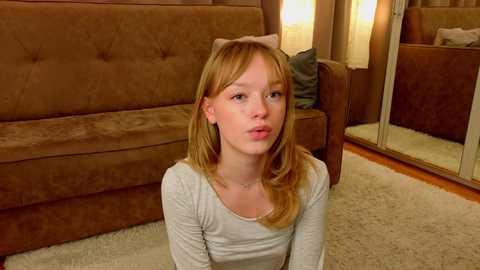 Media: Video of a young Caucasian girl with blonde hair, wearing a light gray top, sitting on a beige carpet in a cozy, dimly lit room with a brown couch, a mirror, and a soft, warm ambiance.