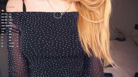 Media: A video of a woman with long, blonde hair wearing a sheer, black top with white polka dots. Her bare shoulder is visible, with a tattoo on her upper arm. The background is blurred, showing a room with soft lighting.