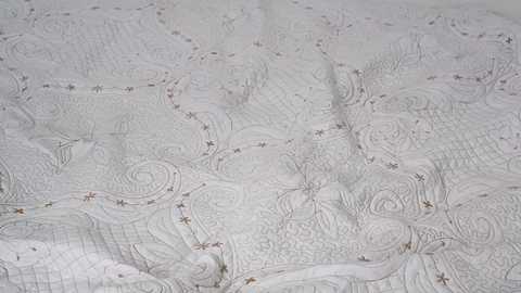 Video of intricate, white quilted fabric with swirling floral patterns and small, scattered star motifs. The fabric texture appears soft and delicate, with detailed stitching creating a textured, quilted appearance.