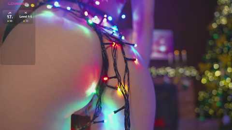 Video of a festive indoor scene featuring a lit-up Christmas tree in the background, with a close-up of a person's bare, white buttocks adorned with colorful string lights. The setting is cozy and holiday-themed.