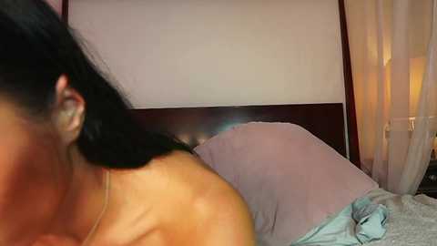 Video of a woman with long black hair, partially visible, lying on a bed with pale sheets and a white curtain in the background.