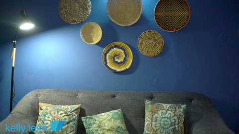 Video of a modern living room with a grey tufted sofa adorned with patterned pillows against a deep blue wall. Above the sofa, a cluster of woven circular wall hangings in earthy tones adds rustic charm. A black floor lamp with a cylindrical shade casts a soft glow.