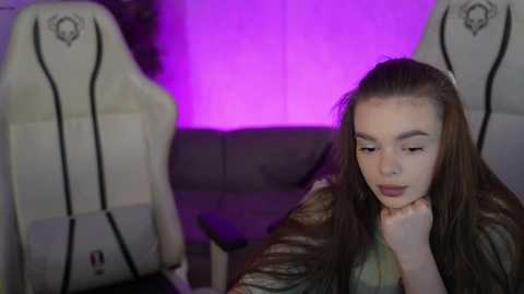 Video of a young Caucasian woman with long brown hair, light skin, and a contemplative expression, seated on a white gaming chair with purple lighting in the background.