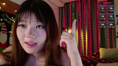 Video of an Asian woman with long, straight brown hair, wearing a black top, pointing at a screen displaying a 'Level 5' achievement in a game. Background features festive Christmas decorations and glowing red and green fairy lights.