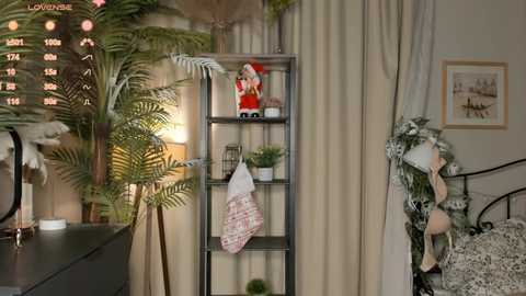 Video of a cozy, Christmas-themed bedroom with a tall, metal shelving unit adorned with a red Santa doll, a white stocking, and potted plants. A faux palm tree stands beside a black metal bedframe with a wreath.