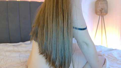 Video of a nude woman with long, straight, light brown hair, sitting on a bed with a gray headboard. She has a blue tattoo on her left arm. Background features a beige wall with a floor lamp.