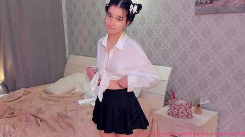 Media: Video of a young Asian woman with dark hair in pigtails, wearing a white shirt tied at the waist and a black skirt, standing in a bedroom with beige bedding, patterned wallpaper, and a nightstand.