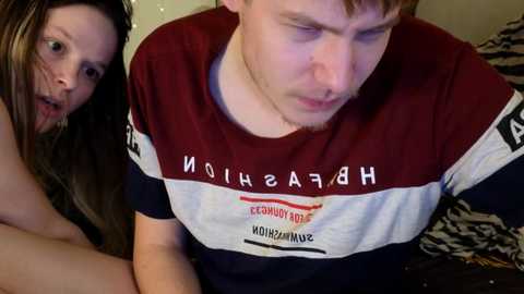 Video of a young man with short red hair, wearing a maroon and white striped shirt, looking down with a contemplative expression. A young woman with long brown hair, wearing a black top, is leaning in closely, appearing concerned.