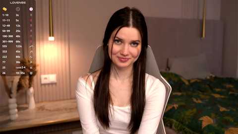Video of a young Caucasian woman with straight, dark brown hair, sitting on a grey chair in a cozy bedroom with a green bedspread and a lit lamp.