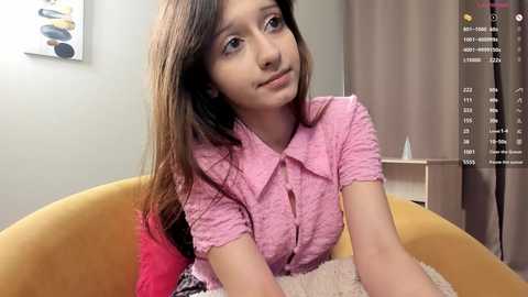 Video of a young girl with long brown hair, wearing a pink button-up shirt, sitting on a mustard-yellow chair. Background includes a beige wall, modern artwork, and a screen displaying a virtual camera interface.