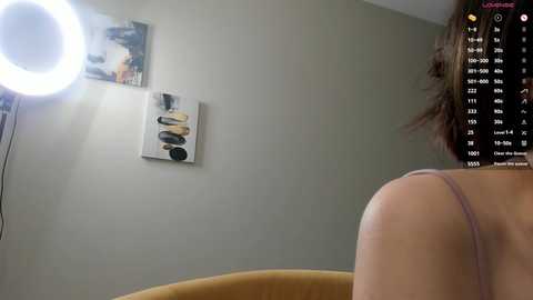 A video of a woman with brown hair in a light pink bra, standing in front of a gray wall with a circular light and abstract art.