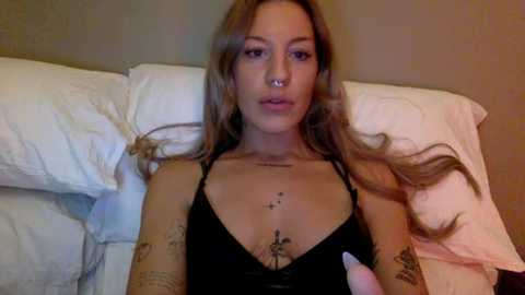 Video of a tattooed young woman with long, wavy brown hair, wearing a black halter top, lying on a bed with white pillows and sheets.
