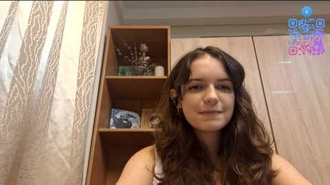 Video of a young woman with long, wavy brown hair, fair skin, and a small nose ring, standing in a bedroom with wooden shelves and lace curtains.