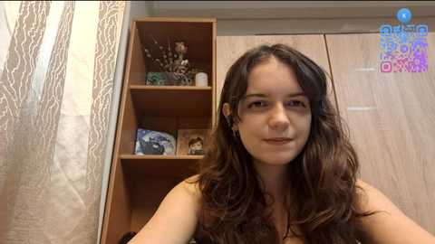 A video of a young woman with long, wavy brown hair, light skin, and a neutral expression, standing in a room with wooden shelves and a mirror.