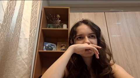 Video of a young woman with long dark hair, wearing a sleeveless top, leaning on her elbow, looking contemplative. Behind her, wooden wardrobes and a shelf with decorative items are visible.