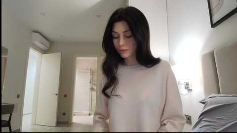 A video of a woman with long black hair, wearing a light beige sweater, standing in a modern, minimalist apartment with white walls and beige furniture.