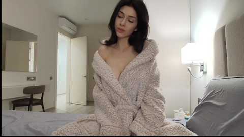 Video of a young woman with fair skin and long black hair, wearing a light beige, textured bathrobe, sitting on a bed in a modern, minimalist bedroom.