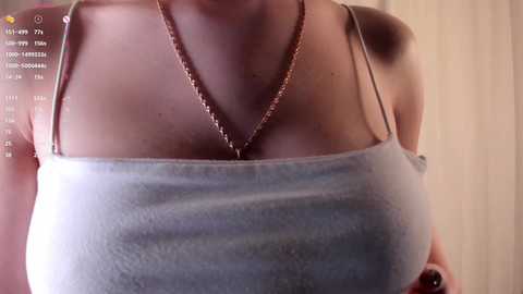 Video of a woman's upper torso, wearing a light blue spaghetti-strap tank top, revealing a gold chain necklace. The background is blurred, possibly a wooden surface.