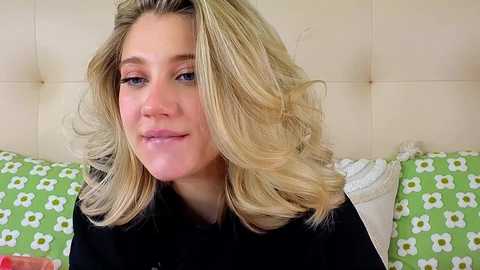 Video of a smiling, blonde woman with wavy hair, wearing a black shirt, against a green floral pillow backdrop.