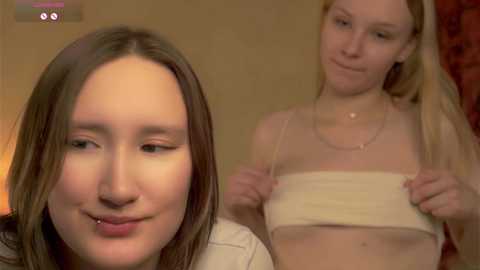 Video of a young woman with straight, light brown hair and light skin, wearing a white tank top, smiling at the camera. Behind her, another woman with blonde hair and a similar tank top adjusts her bra, looking away.