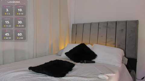 Video of a minimalist bedroom with a gray upholstered headboard, white bedding, black pillow, and a digital clock on the nightstand. A weather forecast overlay shows 3-6 inches of snowfall.