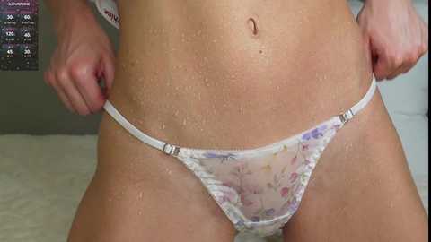 Video of a light-skinned person with a wet, glistening body, wearing semi-transparent floral-patterned panties. The background shows a white bed with a gray wall behind.