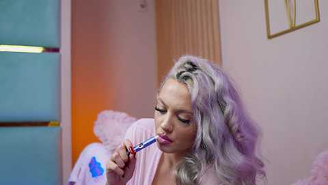 Video of a blonde woman with voluminous curls, wearing a pink robe, using a blue toothbrush in a pastel-colored room with soft lighting.